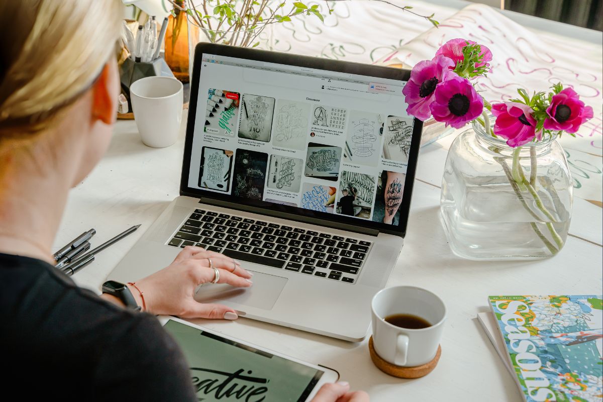 Personne travaillant sur un ordinateur portable et consultant Pinterest, avec une tablette affichant le mot "creative" et une tasse de café à côté. Une ambiance inspirante et créative pour le webdesign.